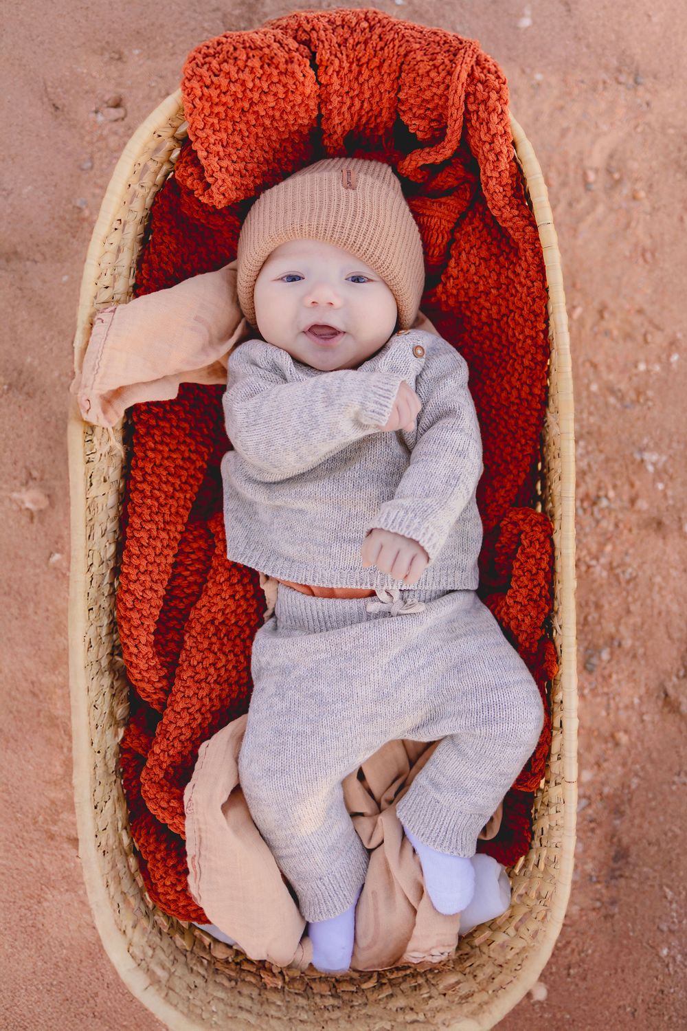 A baby wearing a gray outfit and knit hat lies in a woven basket lined with orange knitted fabric.