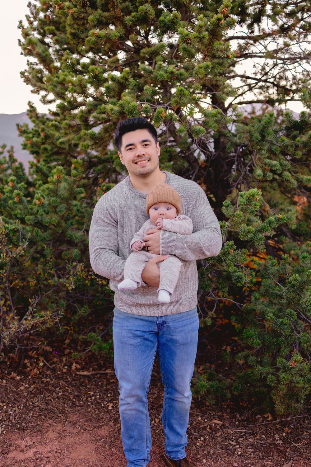 Man in a grey sweater and jeans holds a small baby outdoors in front of an evergreen tree.