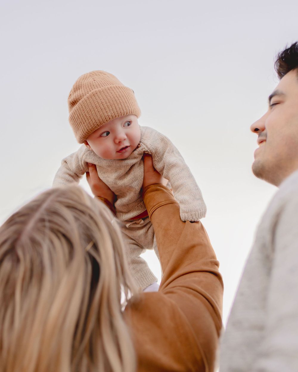 A baby in a beige knit hat is lifted up by someone wearing a brown sweater while another person looks on.