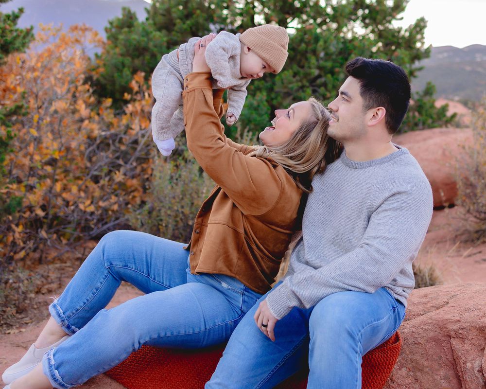 A couple sits together on red rocks during sunset in a desert landscape with autumn foliage visible.  Mom is holding baby boy up in the air