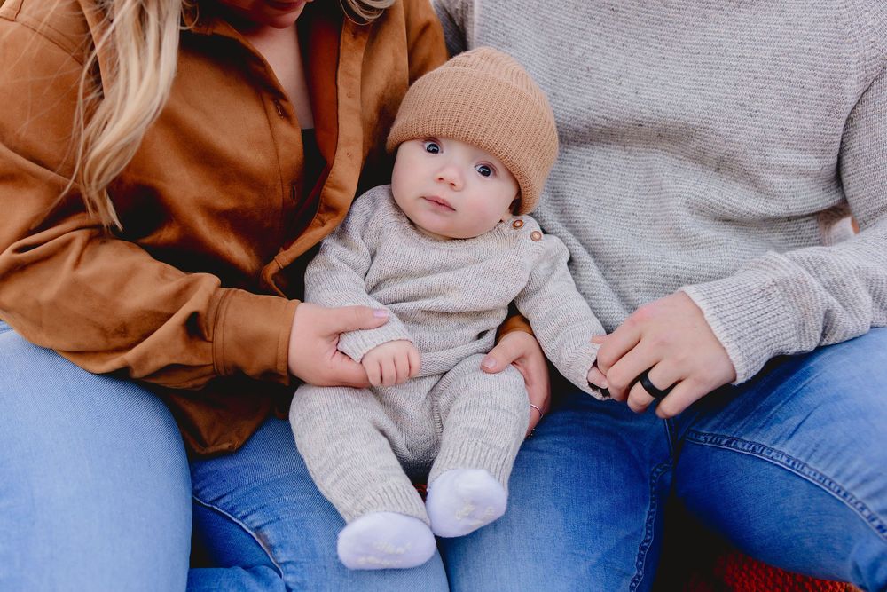A baby wearing a beige knit hat and gray sweater rests on someone's lap in casual denim jeans.