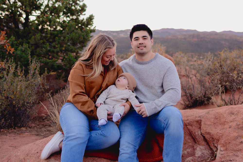 A family sits together on red rocks during sunset in a desert mountain landscape.  Mom, Dad and baby boy