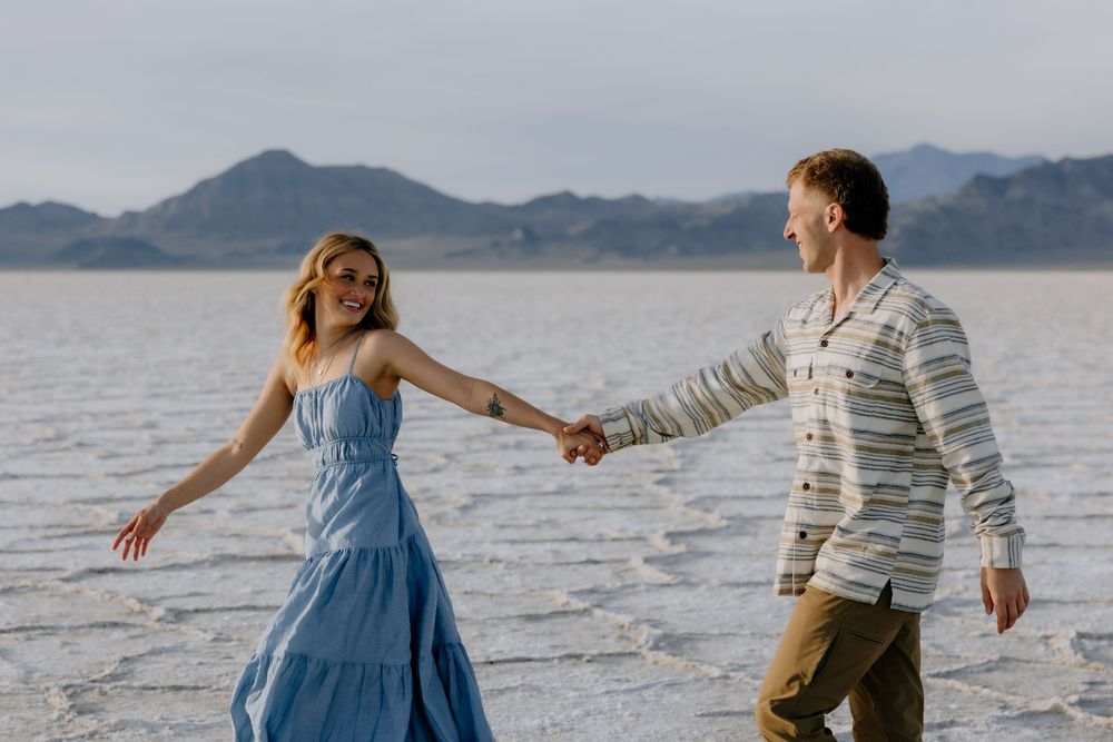 A couple holds hands while walking along the shoreline of a salt lake with mountains in the background.