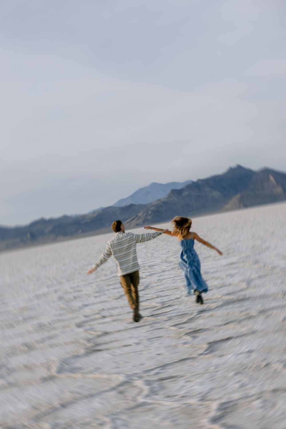 Two people run and leap joyfully across the white salt flats with mountains visible in the misty background.