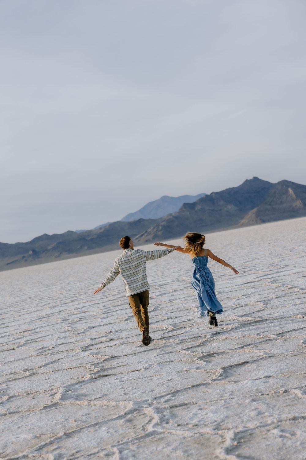 Two people run and dance across the stark white salt flats with mountains visible in the distance.