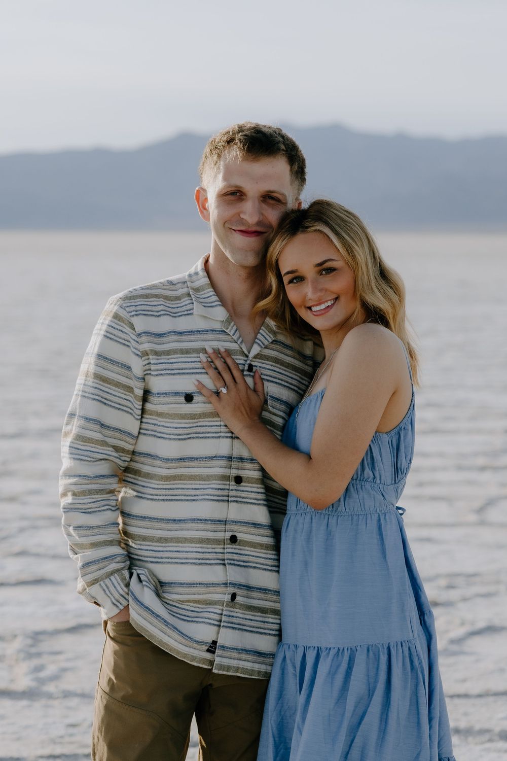 A couple embraces on a beach at sunset with mountains in the background.