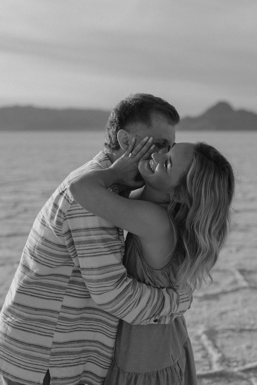 A couple embraces and kisses on a beach at sunset with mountains visible in the background.