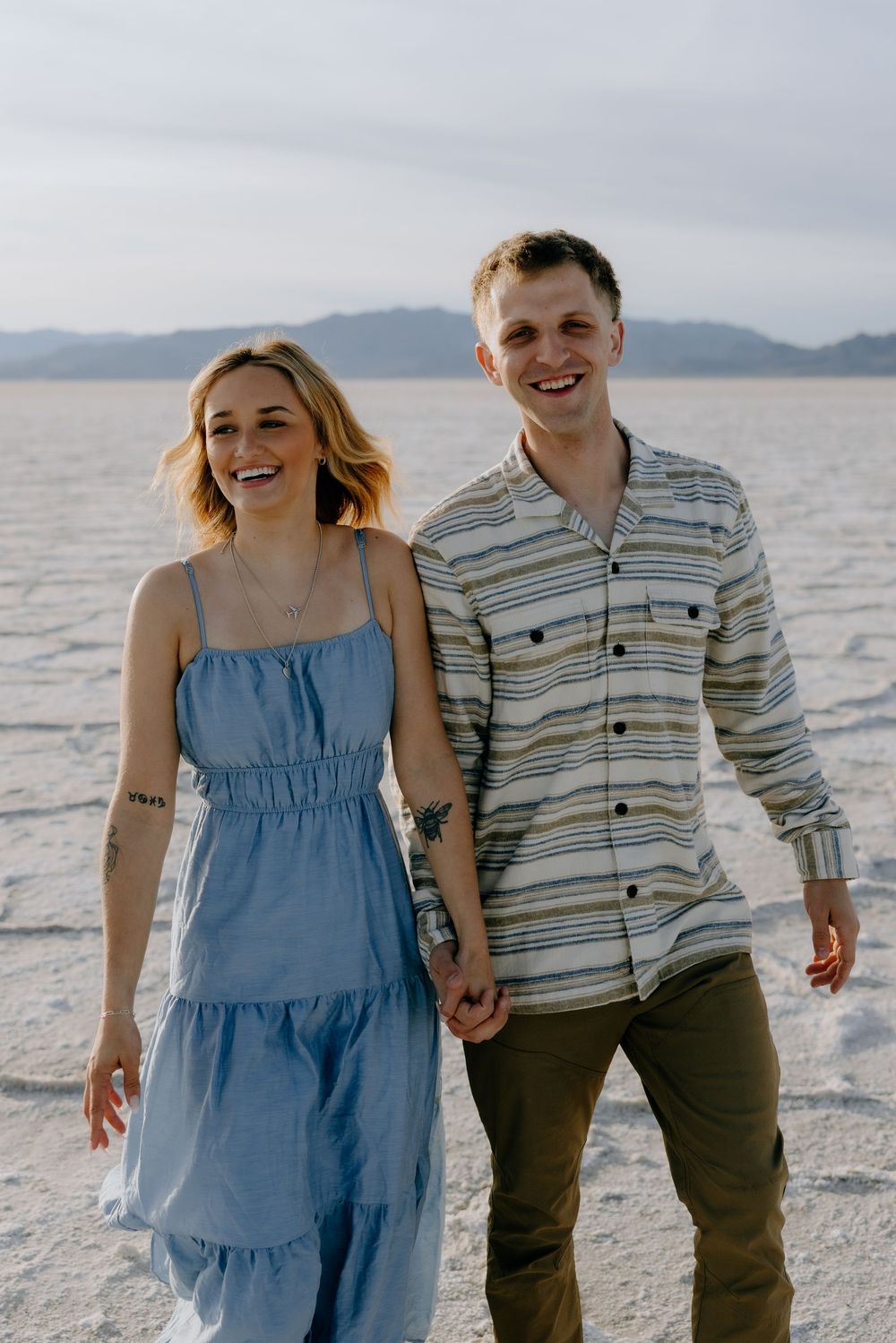 A joyful couple walking and laughing together on white sand with mountains in the background.