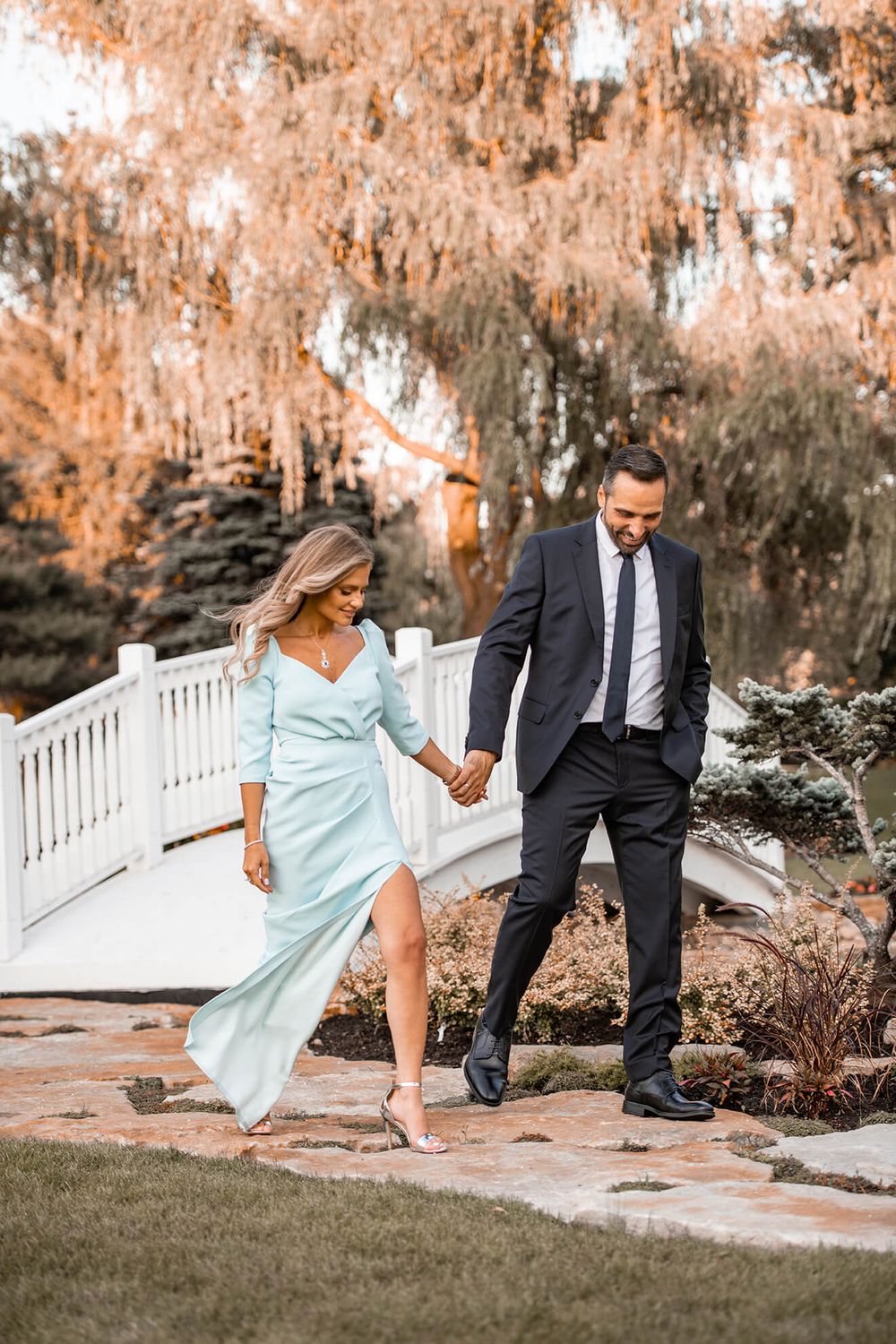 Couple in formal wear playfully walking through garden with white fence.