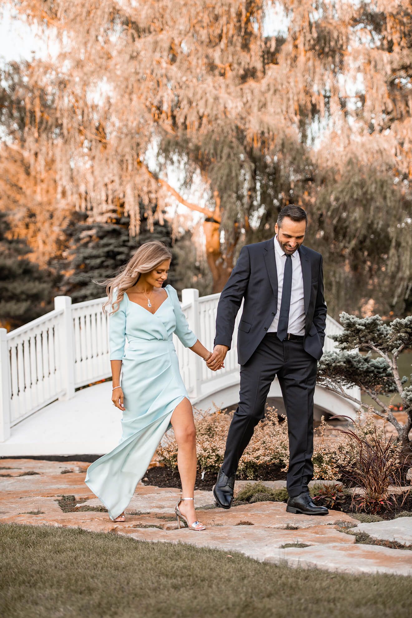 Couple in formal wear playfully walking through garden with white fence.