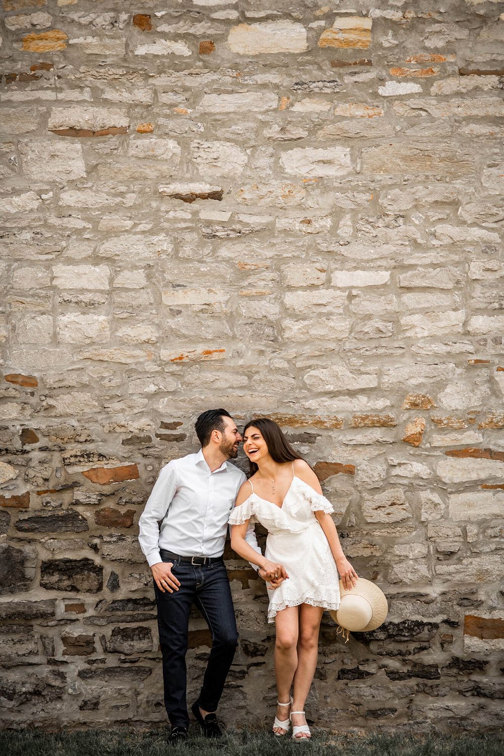Couple in casual attire sitting against rustic stone wall during romantic photoshoot.