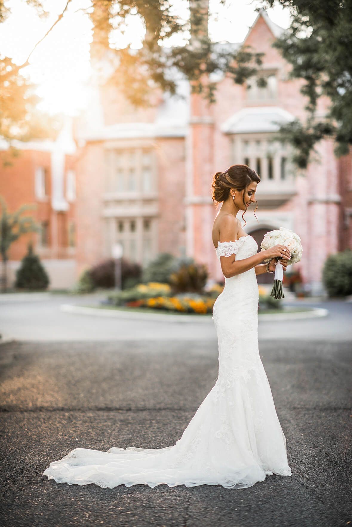 A bride in a flowing white dress poses on a driveway at sunset with a historic building in the background.