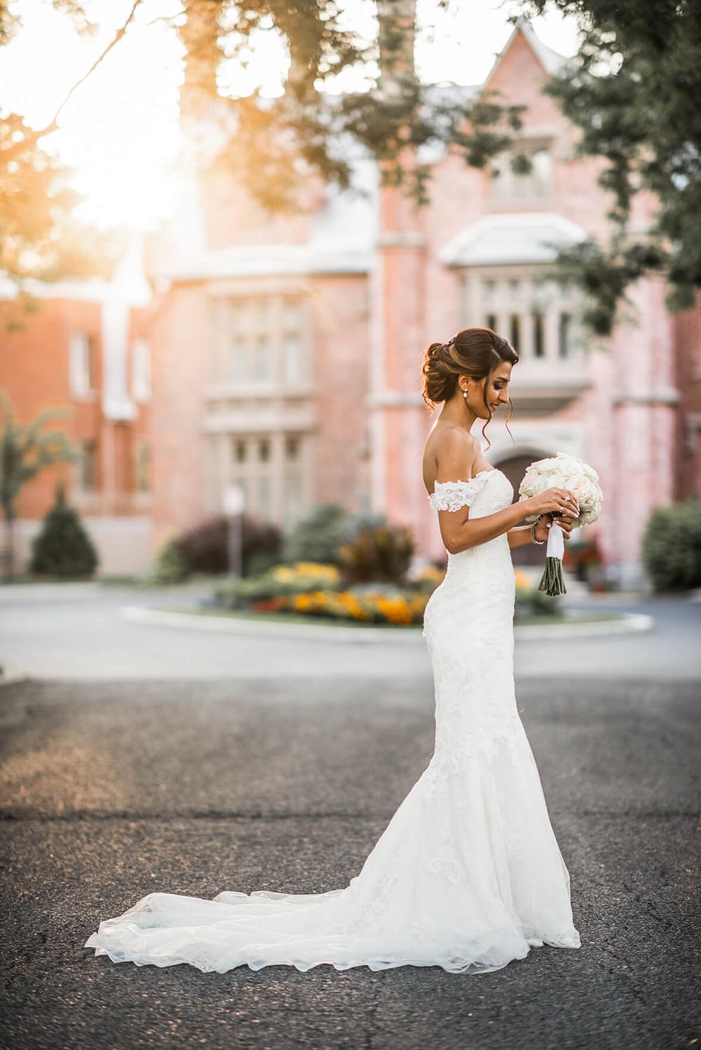 A bride in a flowing white dress poses on a driveway at sunset with a historic building in the background.