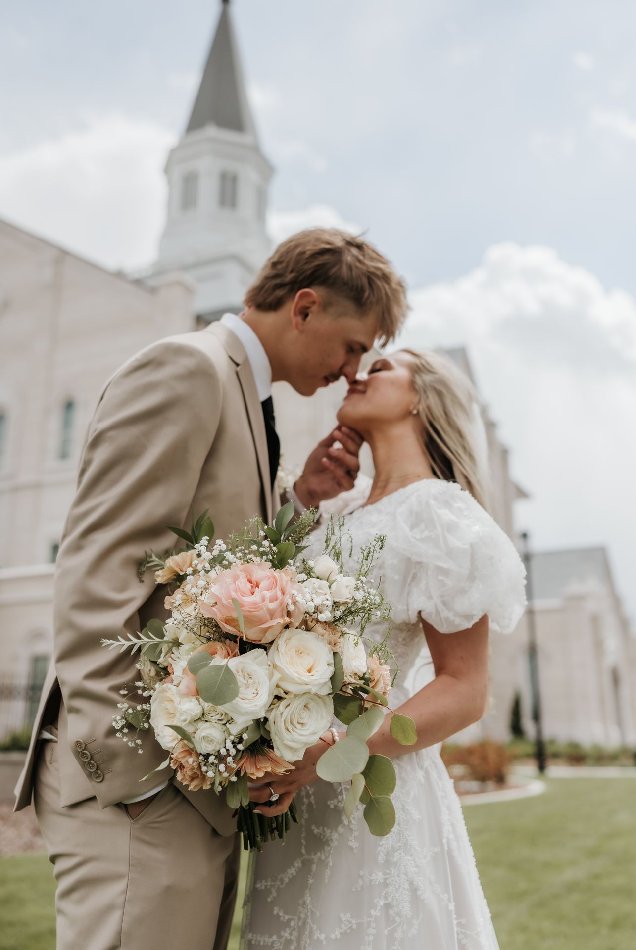 A romantic wedding portrait of a couple sharing a tender moment in front of lds taylorsville temple with a bouquet of roses.