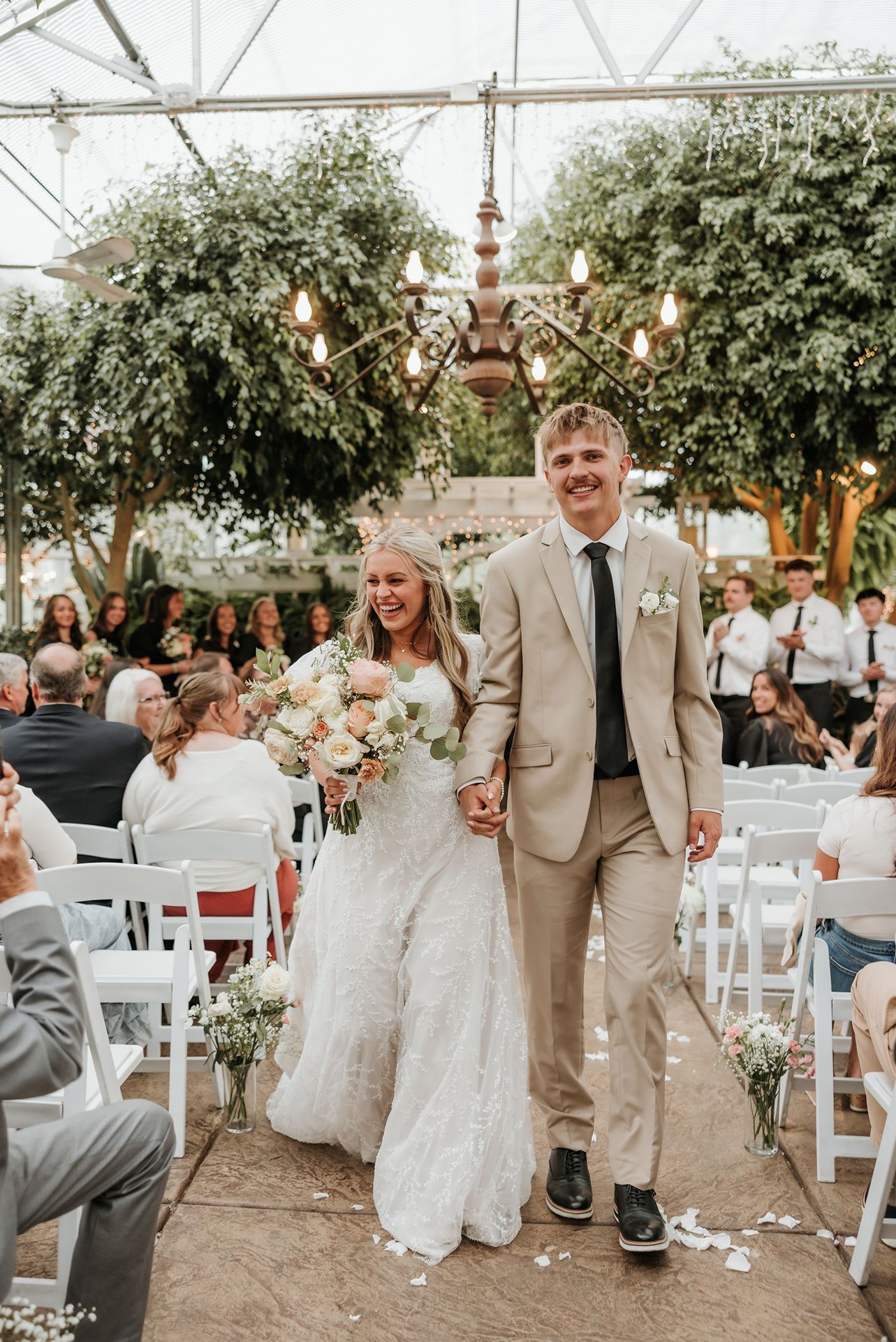 Le Jardin Newlyweds walking down the aisle under a greenhouse tent with hanging lights and white chairs.