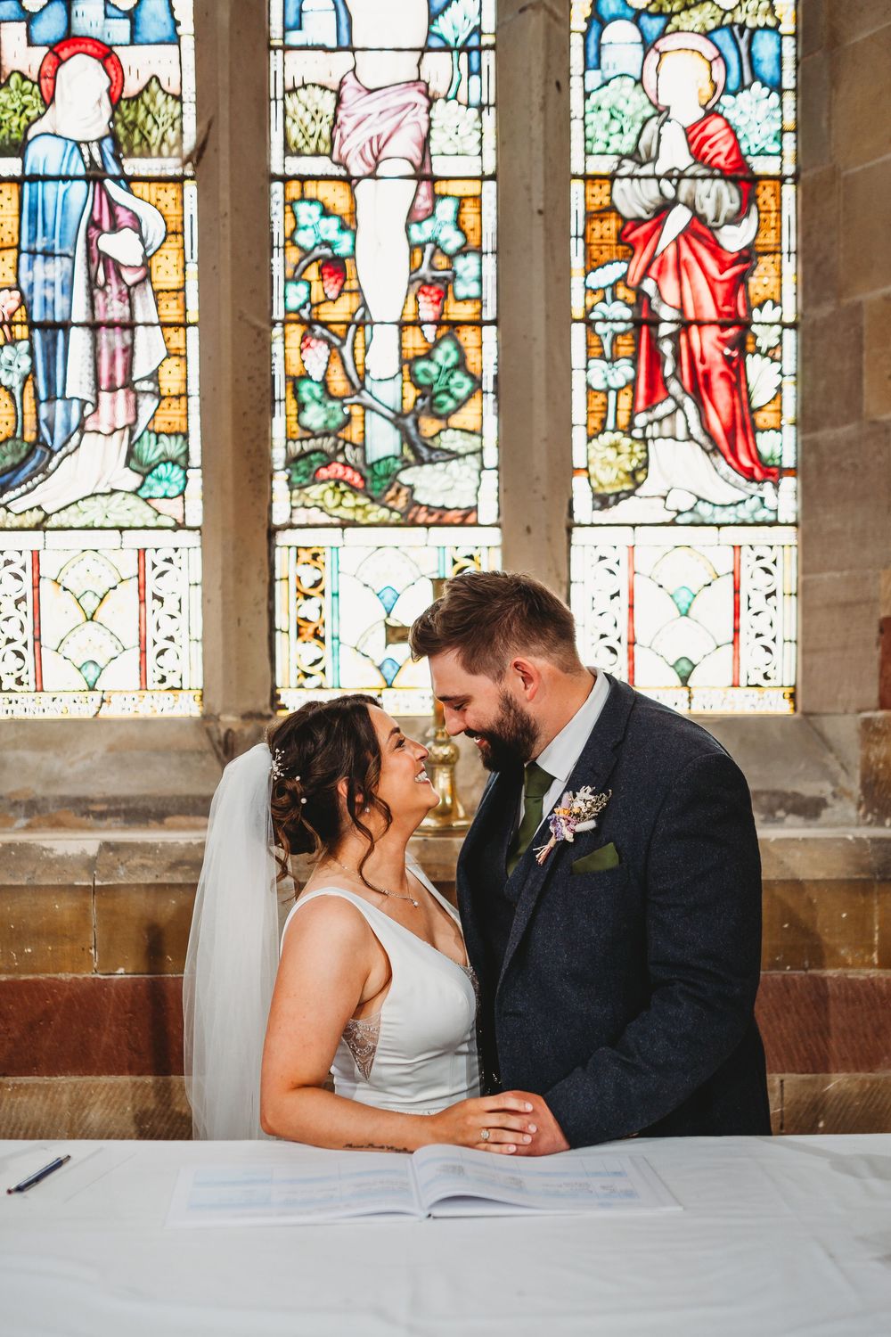 Wedding couple signing documents at a table beneath beautiful stained glass church windows.