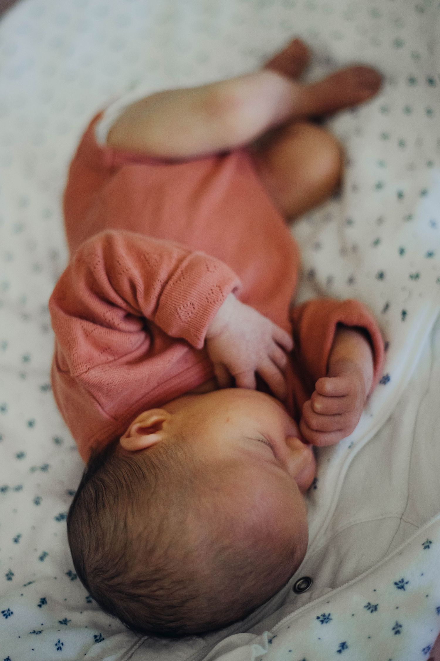 Newborn baby in orange outfit lying on a floral white blanket, eyes closed peacefully.