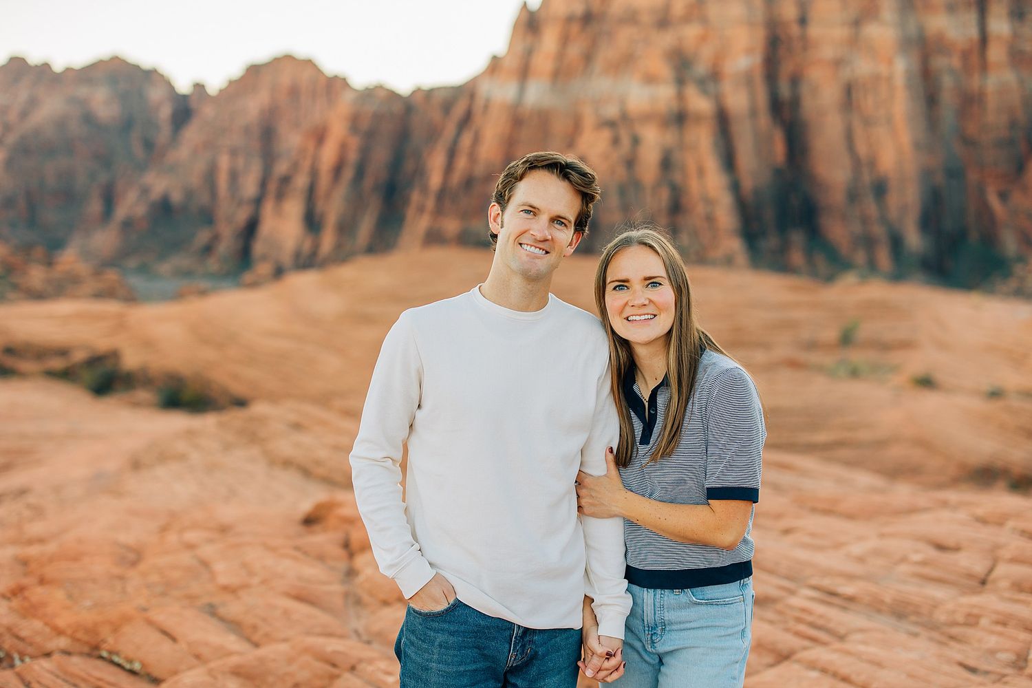 A couple poses together on red rock formations in a desert landscape during sunset.