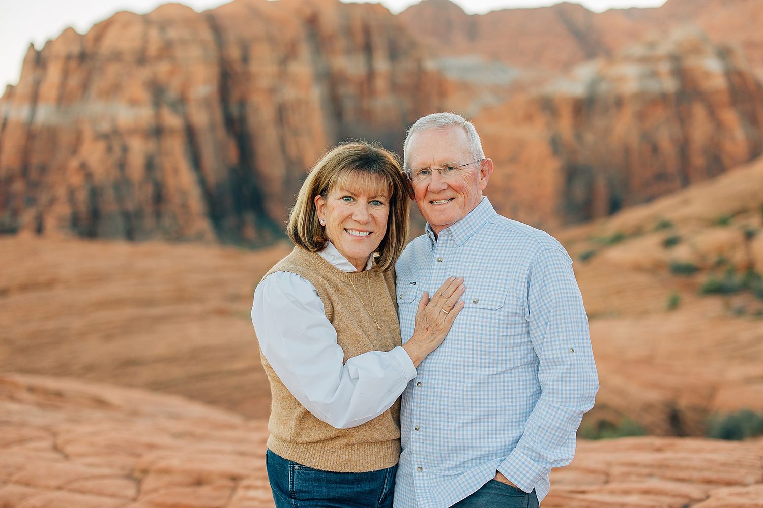 Couple in casual attire share tender moments against backdrop of majestic desert cliffs at sunset.