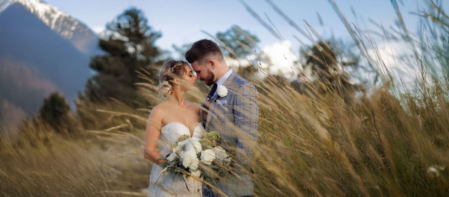 Romantic bride and groom embracing in tall golden grass with stunning mountain backdrop at sunset.