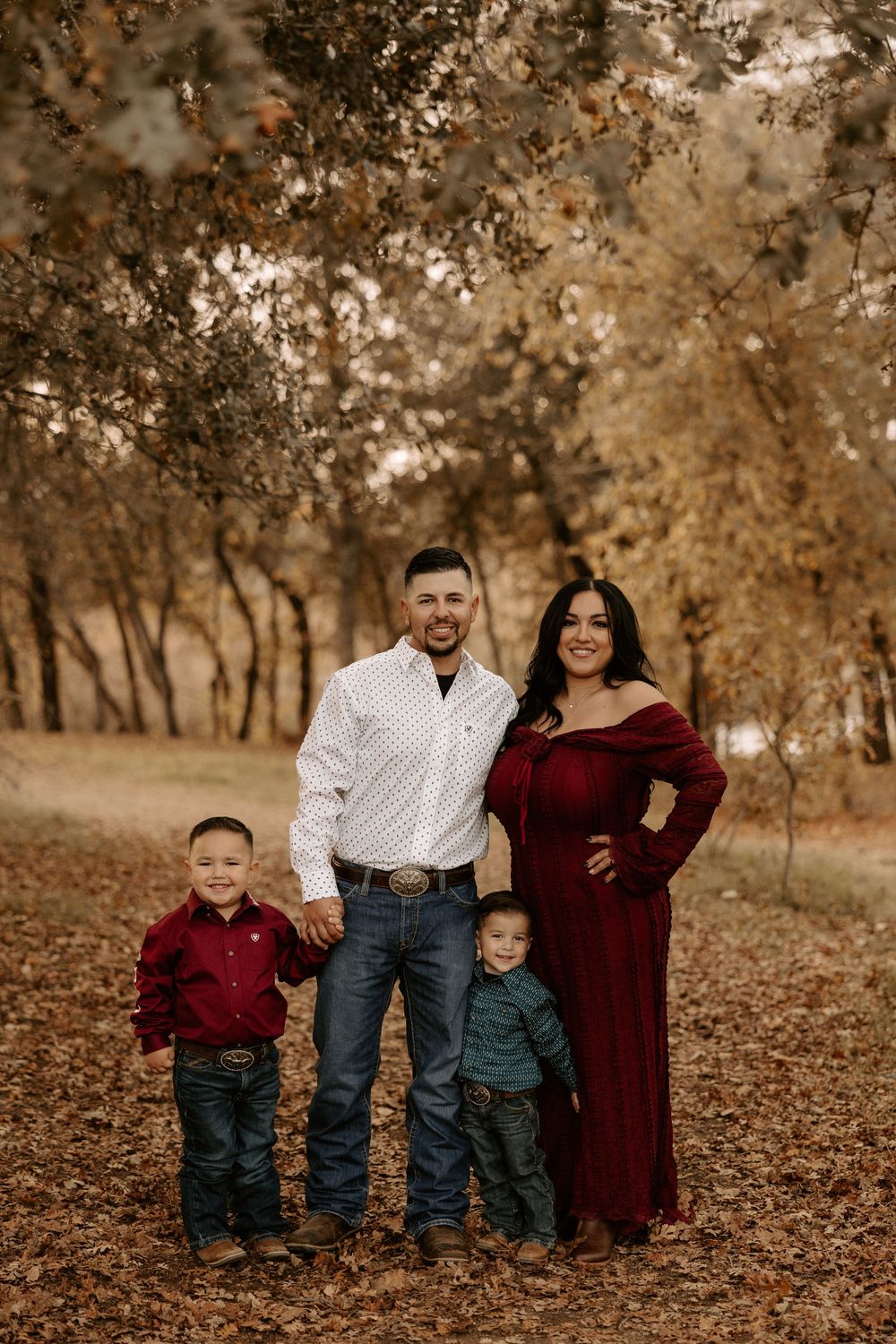 A family in coordinating burgundy and white outfits poses together on fallen autumn leaves in a wooded setting.