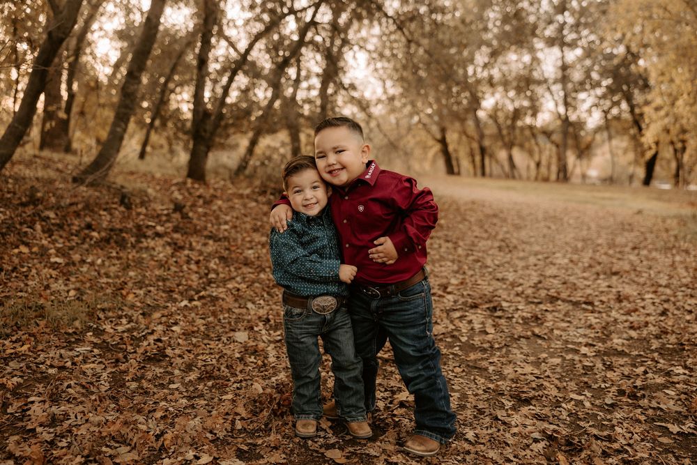 Two young children in denim and red jackets play together in a park filled with fallen autumn leaves.