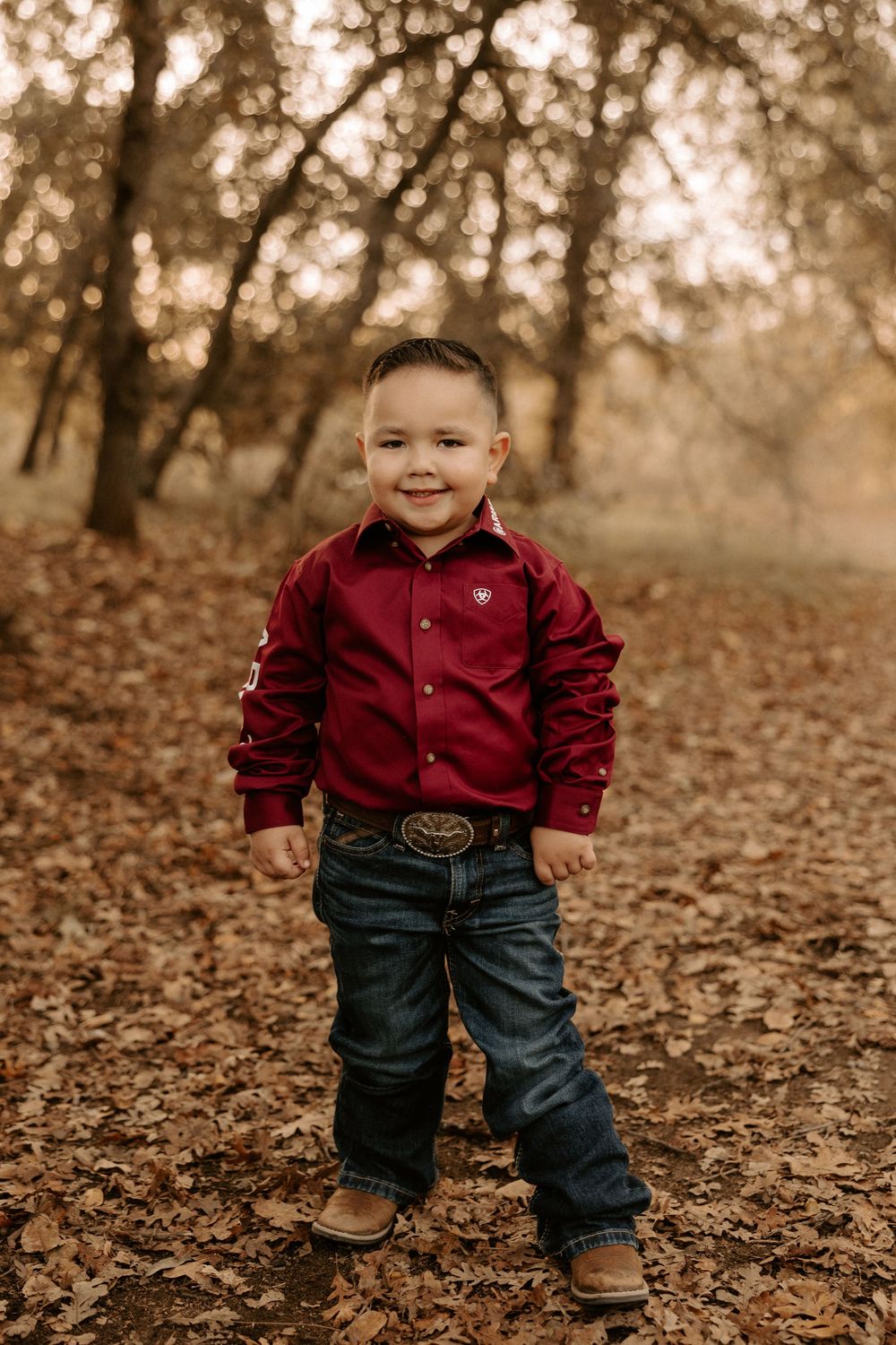 Young child in red shirt and jeans stands playfully among fallen autumn leaves in a wooded setting.