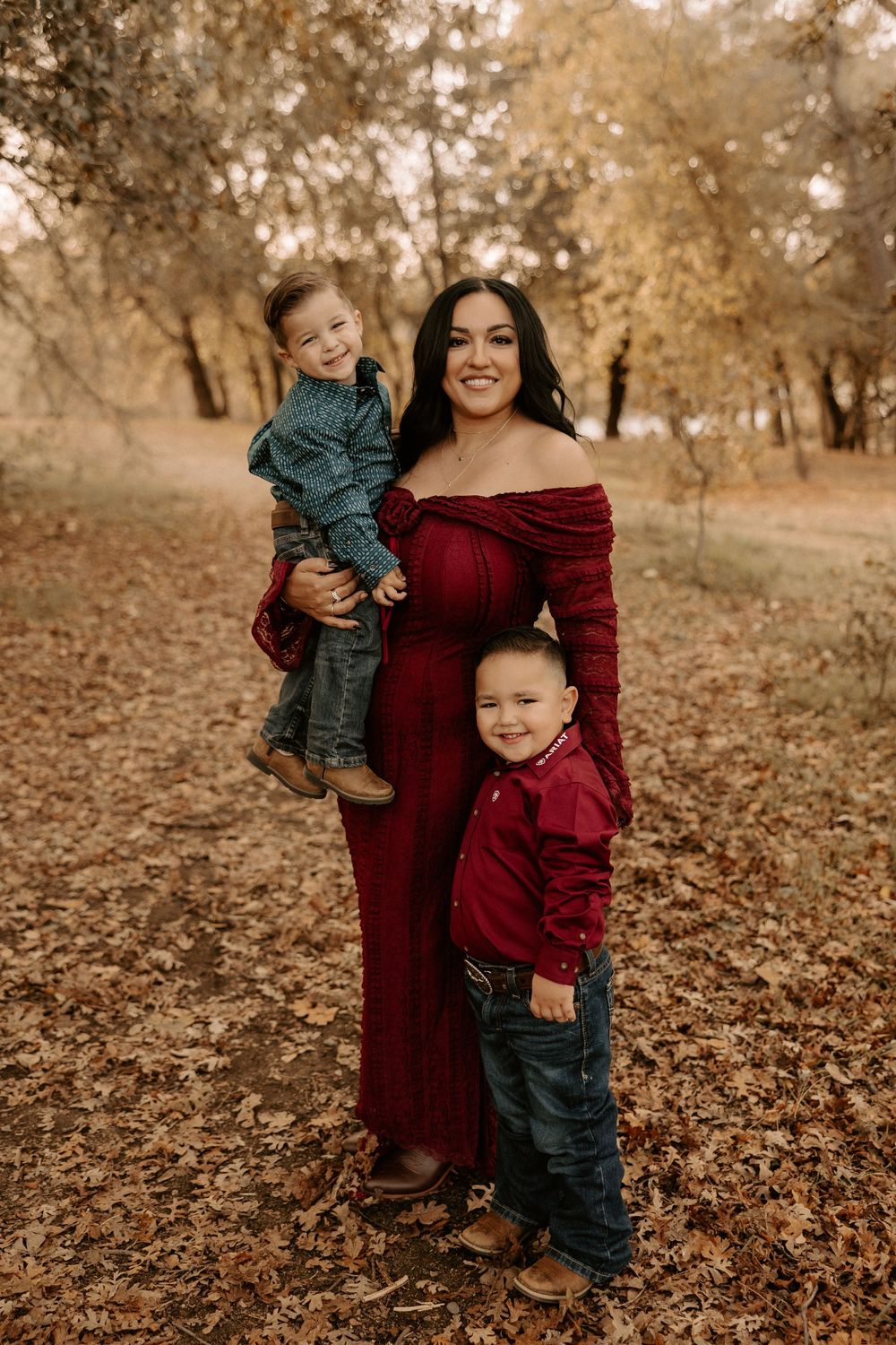 A family wearing burgundy and denim poses together on fallen autumn leaves in a wooded setting during golden hour.