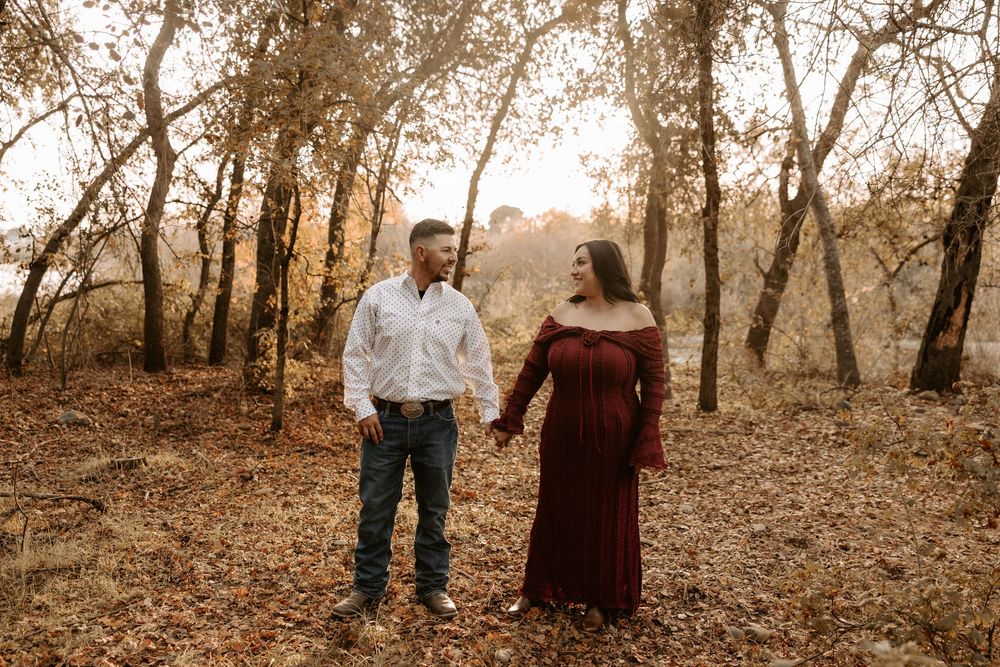 A couple stands together in a wooded area during autumn with golden sunlight filtering through the trees.
