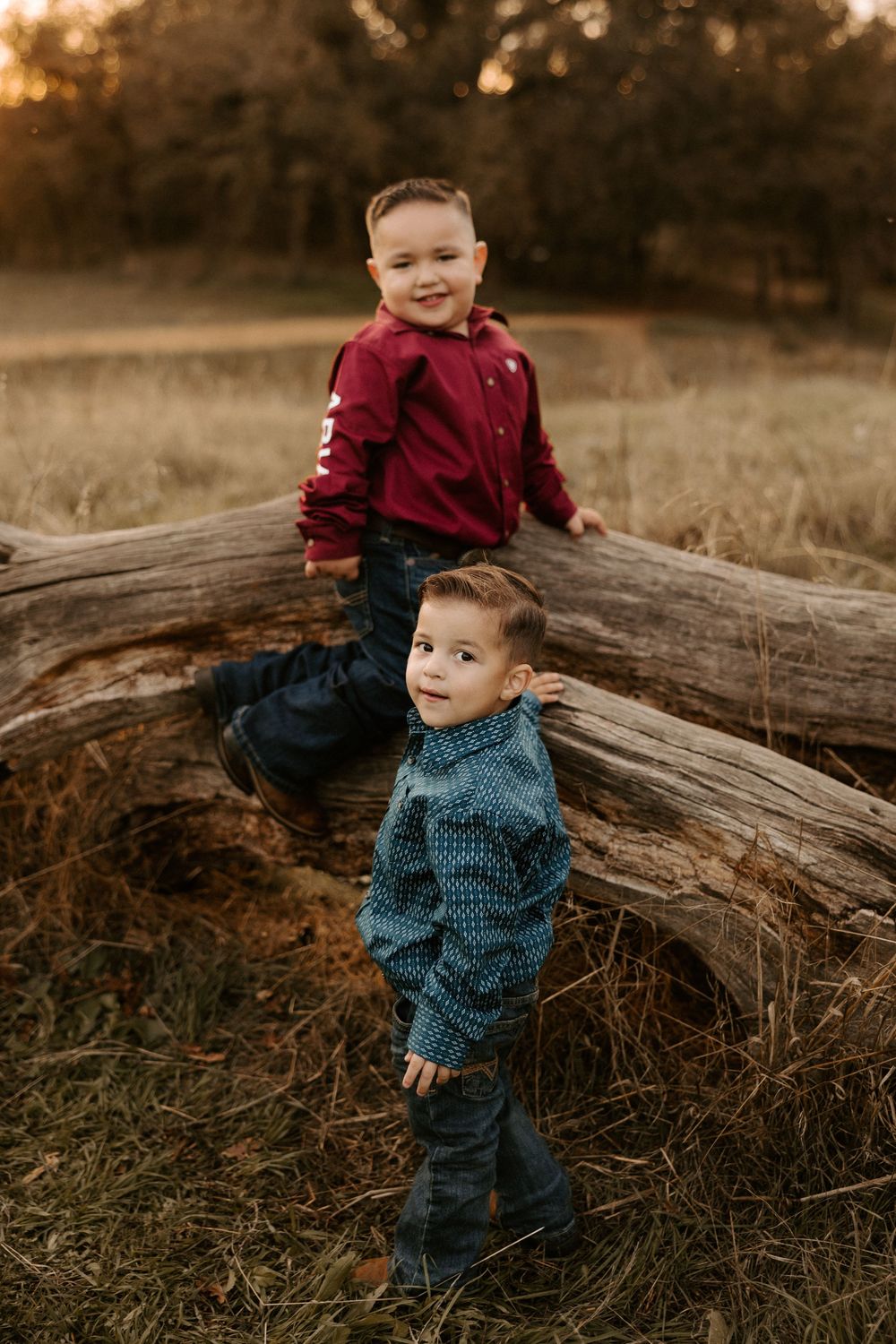 A child in burgundy and navy outfits plays on a fallen log during golden hour in a rustic outdoor setting.