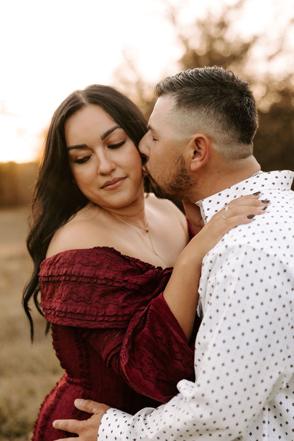Romantic couple embraces at sunset, with woman in burgundy dress and man in white polka dot shirt.