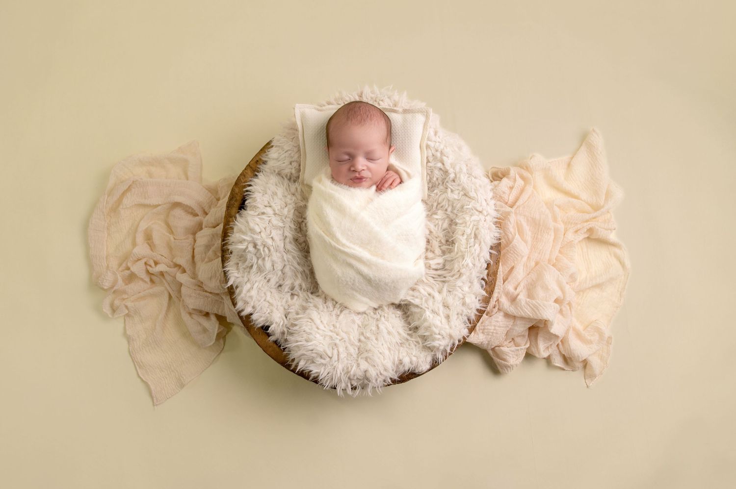 A sleeping newborn baby wrapped in soft white lace fabric lies peacefully on a beige backdrop.