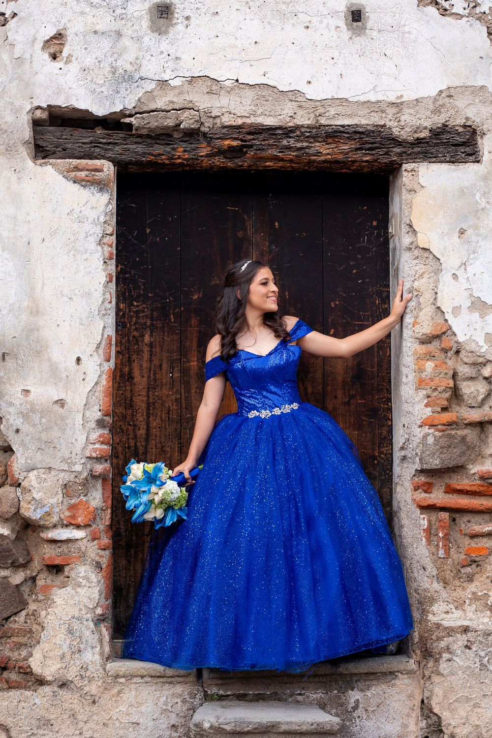 een wearing blue quinceañera dress standing in rustic doorway in Antigua Guatemala holding bouquet