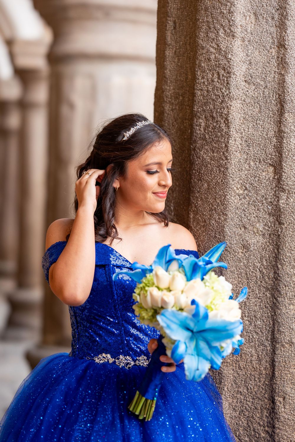 Quinceañera in blue gown holding bouquet beside stone columns in historic building in Antigua Guatemala.