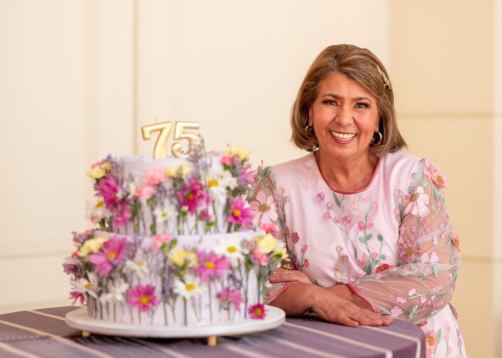 Woman smiling next to floral birthday cake celebrating 75th birthday indoors.