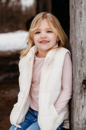 Child wearing a white puffy vest and pink shirt poses playfully against wooden posts in winter setting.