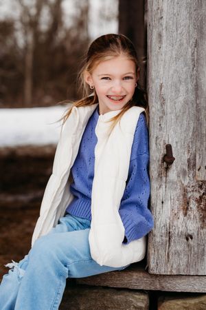 Child in blue dress and white vest sits on rustic wooden structure outdoors during winter.