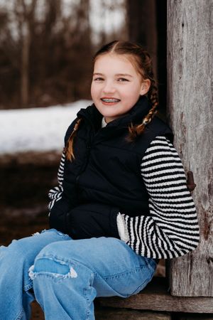 A person wearing a striped shirt and jeans sits casually on a wooden fence in a winter setting.