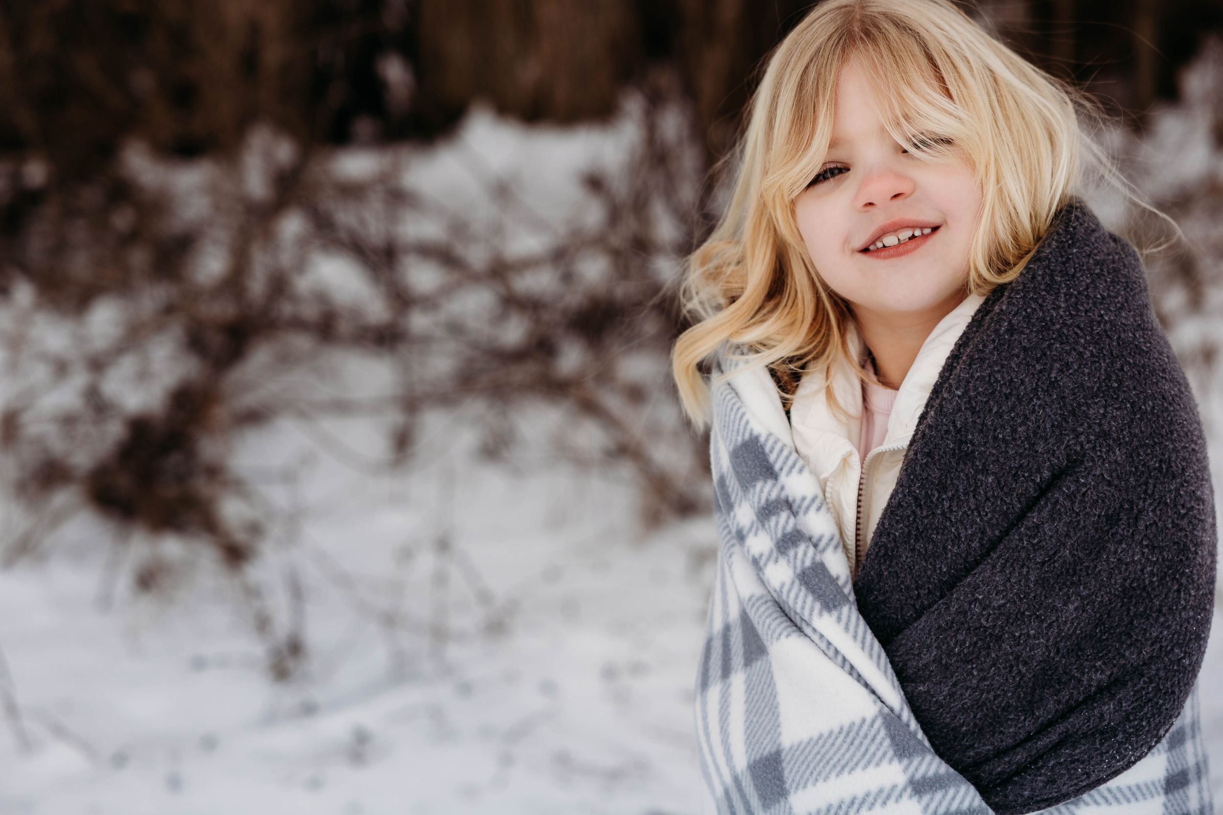 A child wearing a cozy gray sweater and plaid scarf smiles while playing in a snowy winter setting.