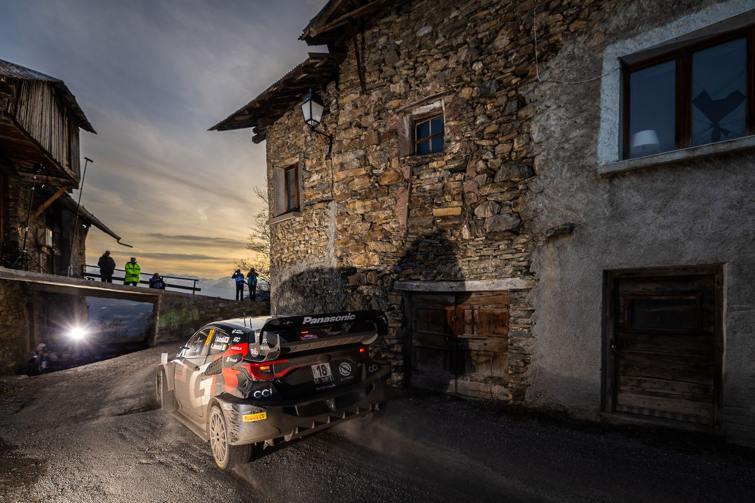 Rally car races along a winding mountain road with snow-capped peaks and rugged cliffs in the background.