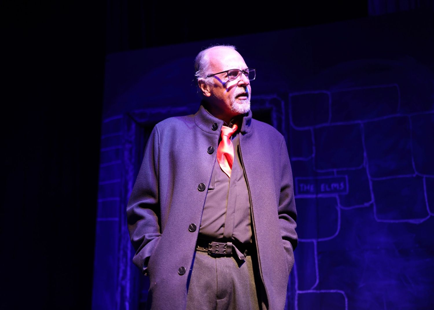 A performer in a gray suit gestures dramatically on a dark stage illuminated by blue lighting.