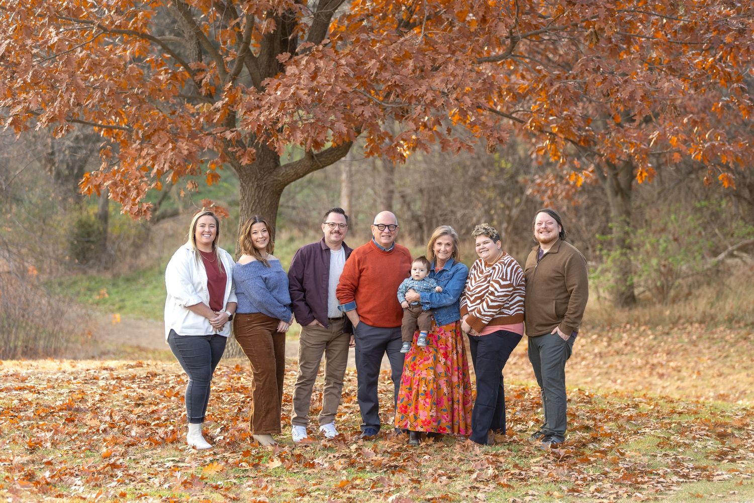 A group stands together outdoors under autumn trees with orange foliage and fallen leaves on the ground.
