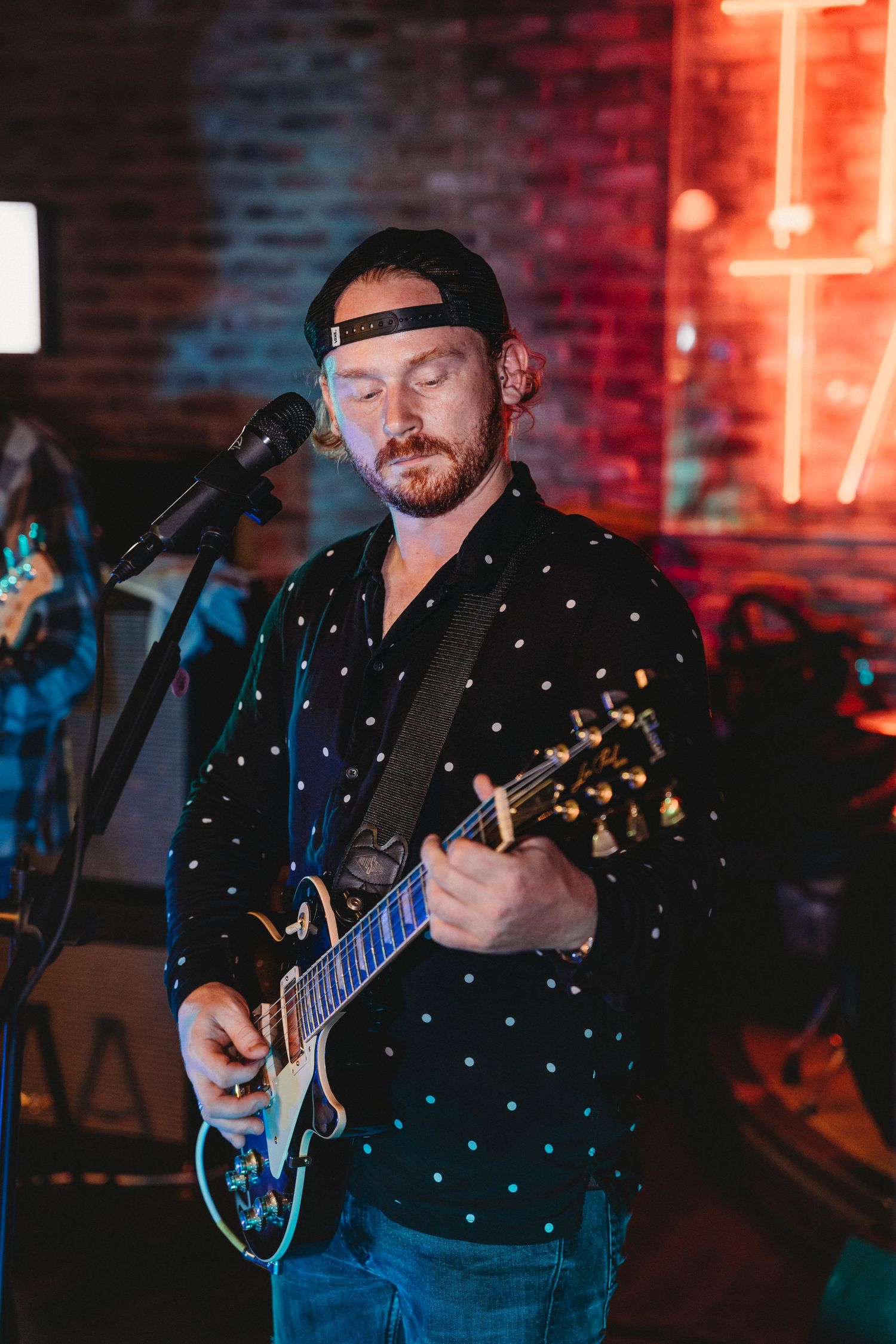 Musician performs on stage with electric guitar wearing polka dot shirt against red and blue lighting backdrop.