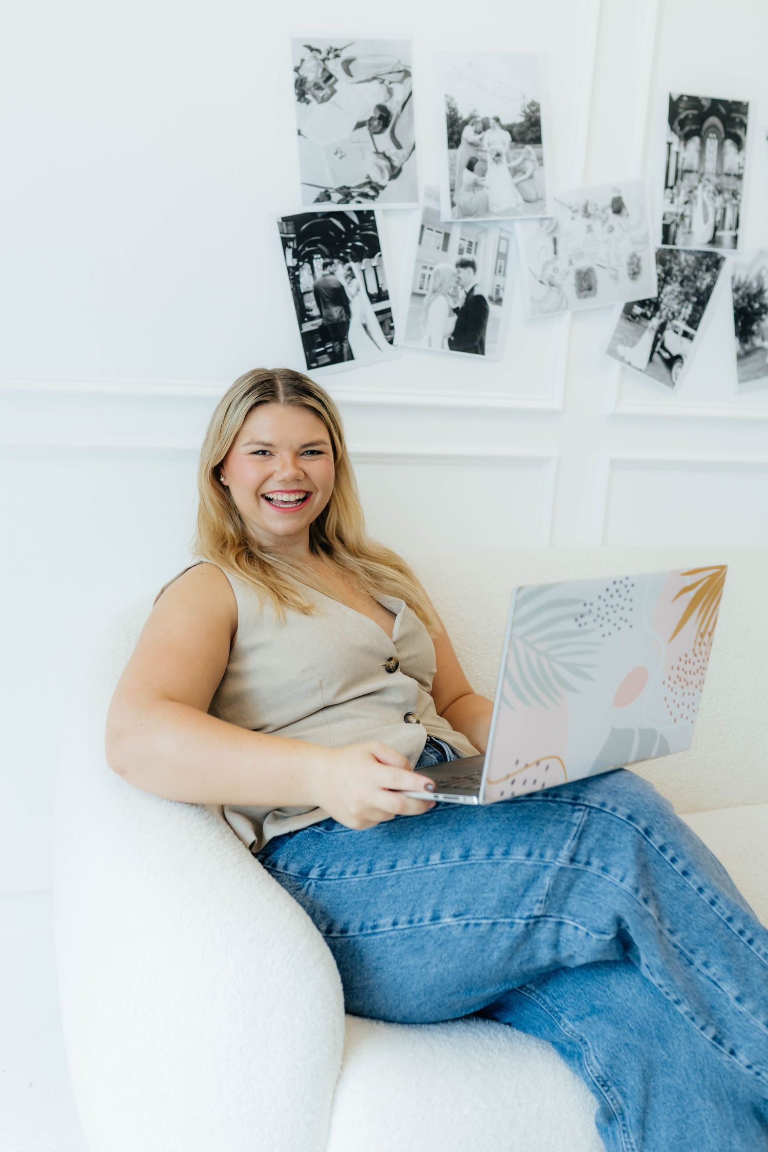 Person in beige tank top and jeans sits comfortably on white couch while working on laptop with photos displayed on wall.