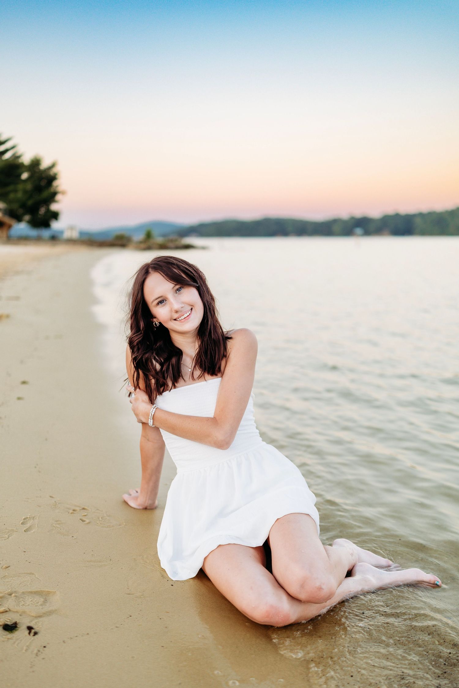 Young woman in white dress sitting on sandy beach shore at sunset with calm water behind her.