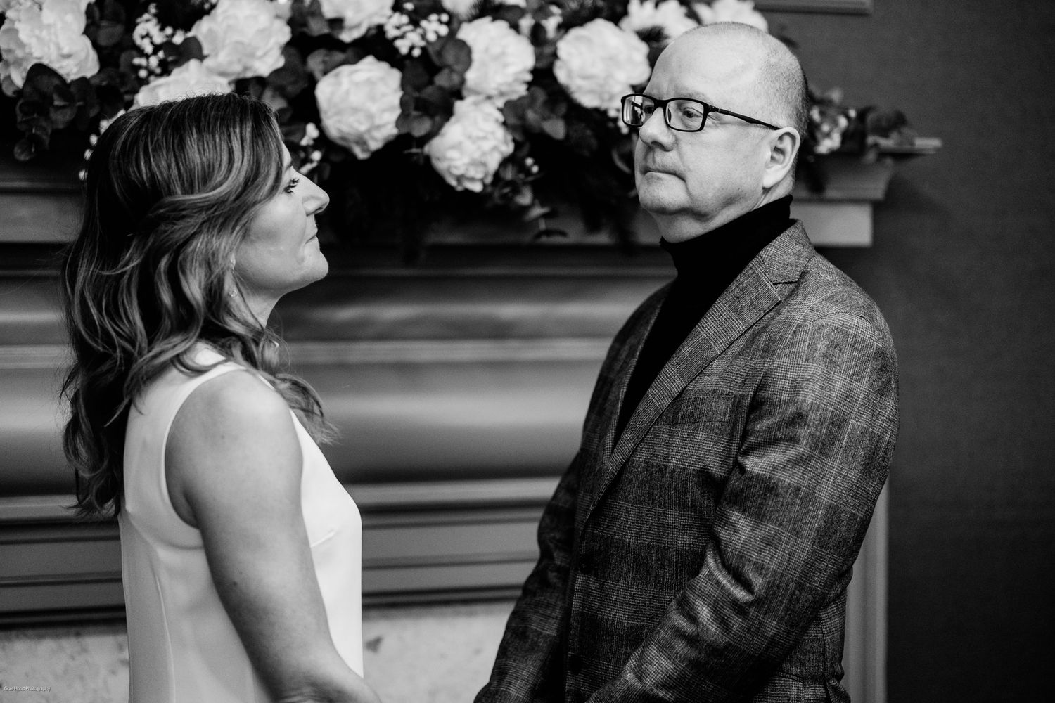 A black and white candid moment between two people in formal attire with floral decorations in the background.
