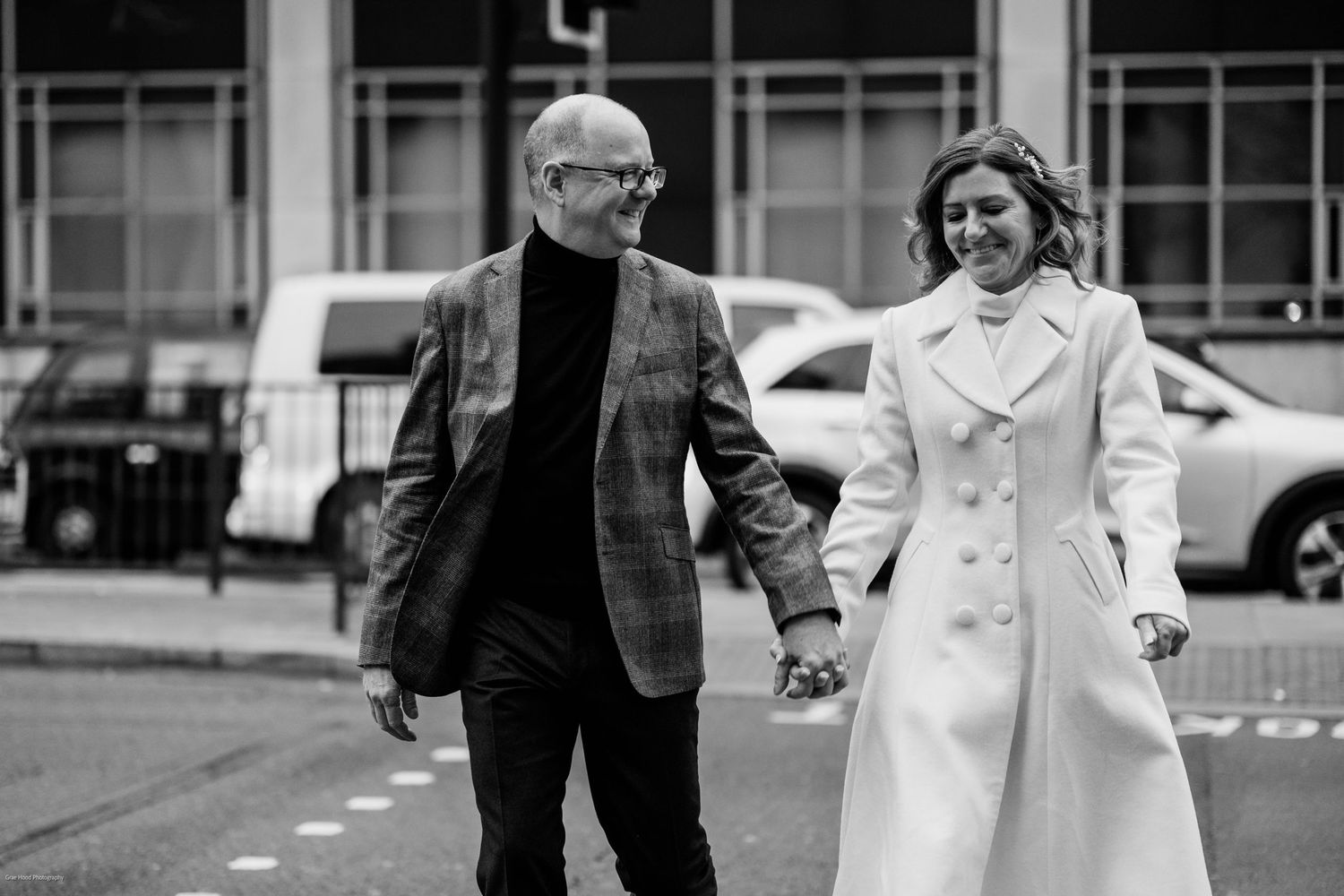 A black and white photo shows a couple walking together outdoors, with the woman wearing a light-colored coat.