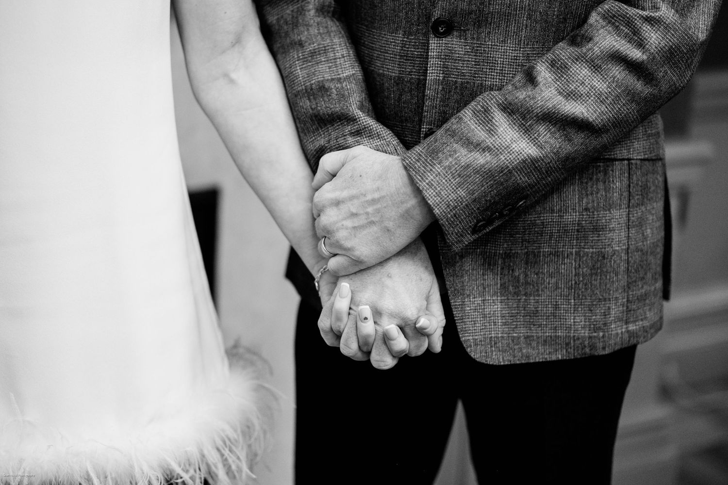 Close-up black and white photo showing two people holding hands with wedding rings visible.