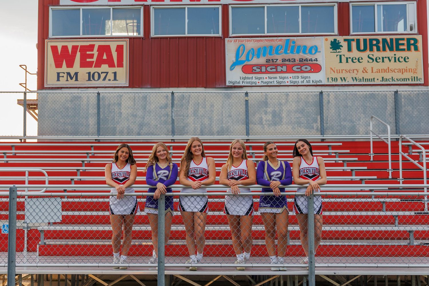 Red and white bleachers at an outdoor sports stadium with blue benches and advertising signage visible.