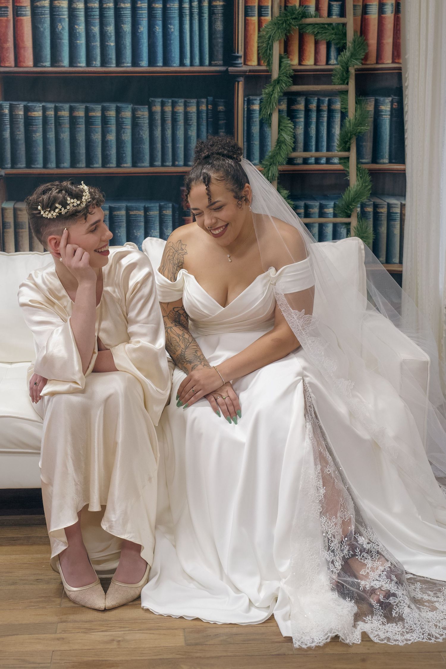 Two people in white wedding attire share an intimate moment while sitting together in front of a bookshelf.